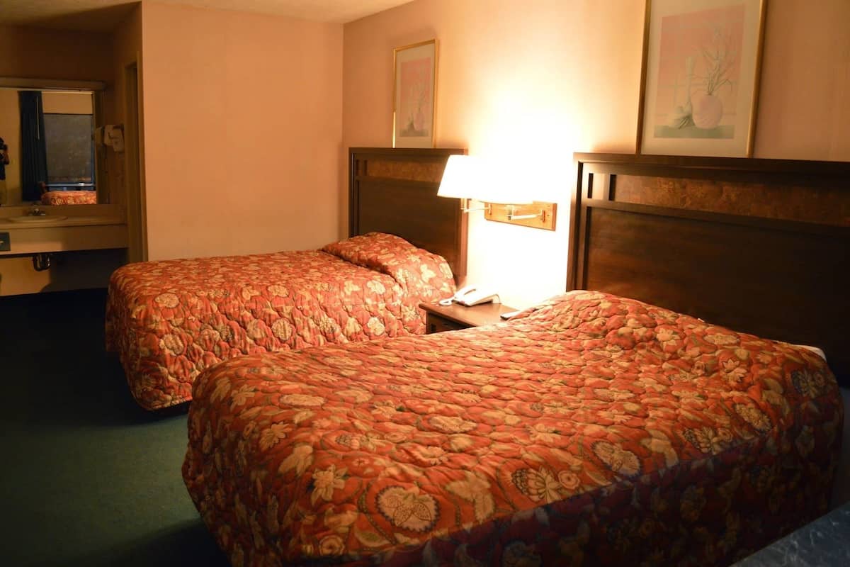 A hotel room with two beds, a bedside table with a lamp, a mirror, and framed artwork on the wall.