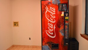 Un distributeur automatique de Coca-Cola, placé dans un coin, avec une petite étagère et une fenêtre à proximité.