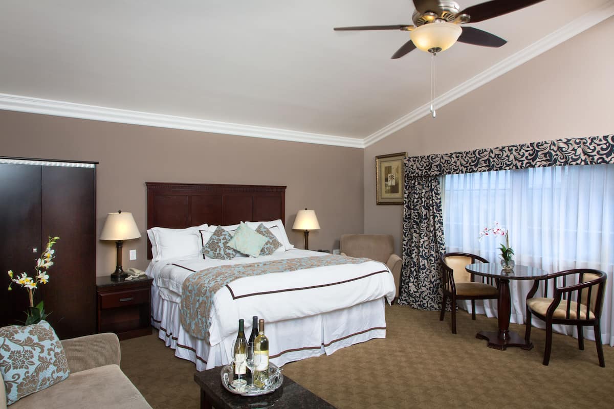 A hotel room with a bed, two chairs, a table with a vase, and a ceiling fan.