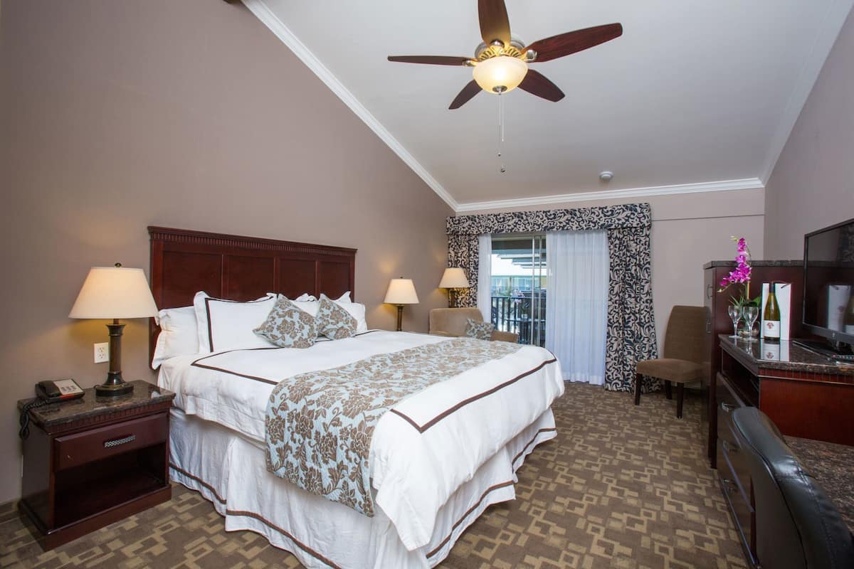 A hotel room with a large bed, two bedside lamps, a ceiling fan, a TV, a minibar, and a balcony with curtains.