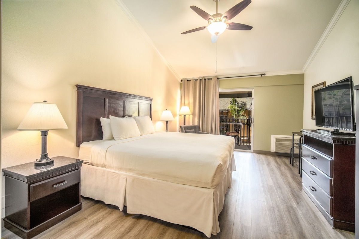 A hotel room with a bed, bedside lamps, a ceiling fan, a TV, and a desk with drawers.