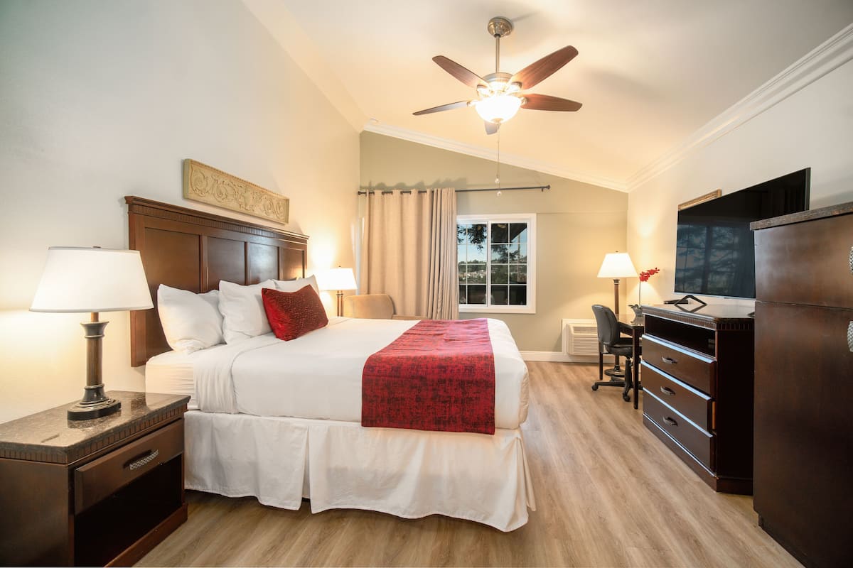 A hotel room with a bed, bedside lamps, a desk with a chair, a television, and a ceiling fan.