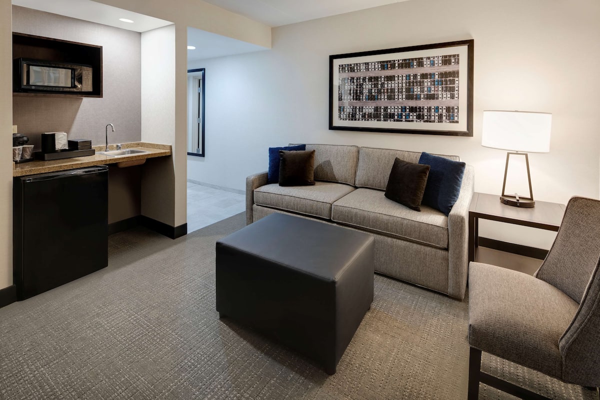 A modern hotel room with a kitchenette, a grey sofa, a black ottoman, and a framed abstract artwork on the wall.