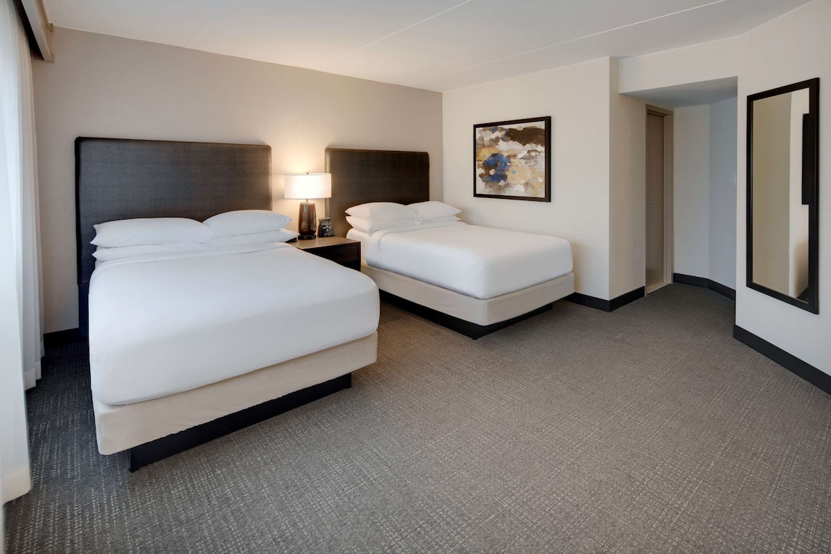 A hotel room with two beds, a nightstand with a lamp, a painting on the wall, and a mirror.