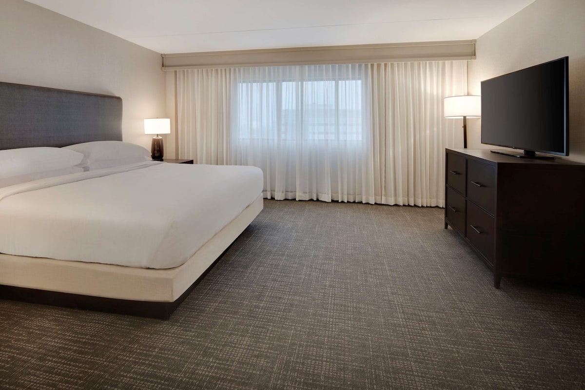 A hotel room with a large bed, a flat-screen TV, a nightstand with a lamp, and a large window with sheer curtains.