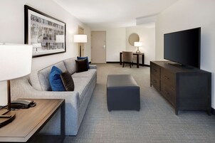 Una habitación de hotel moderna con un sofá gris, un televisor de pantalla plana, una mesita de madera, una pequeña mesa redonda con una lámpara y un cuadro grande enmarcado en la pared.
