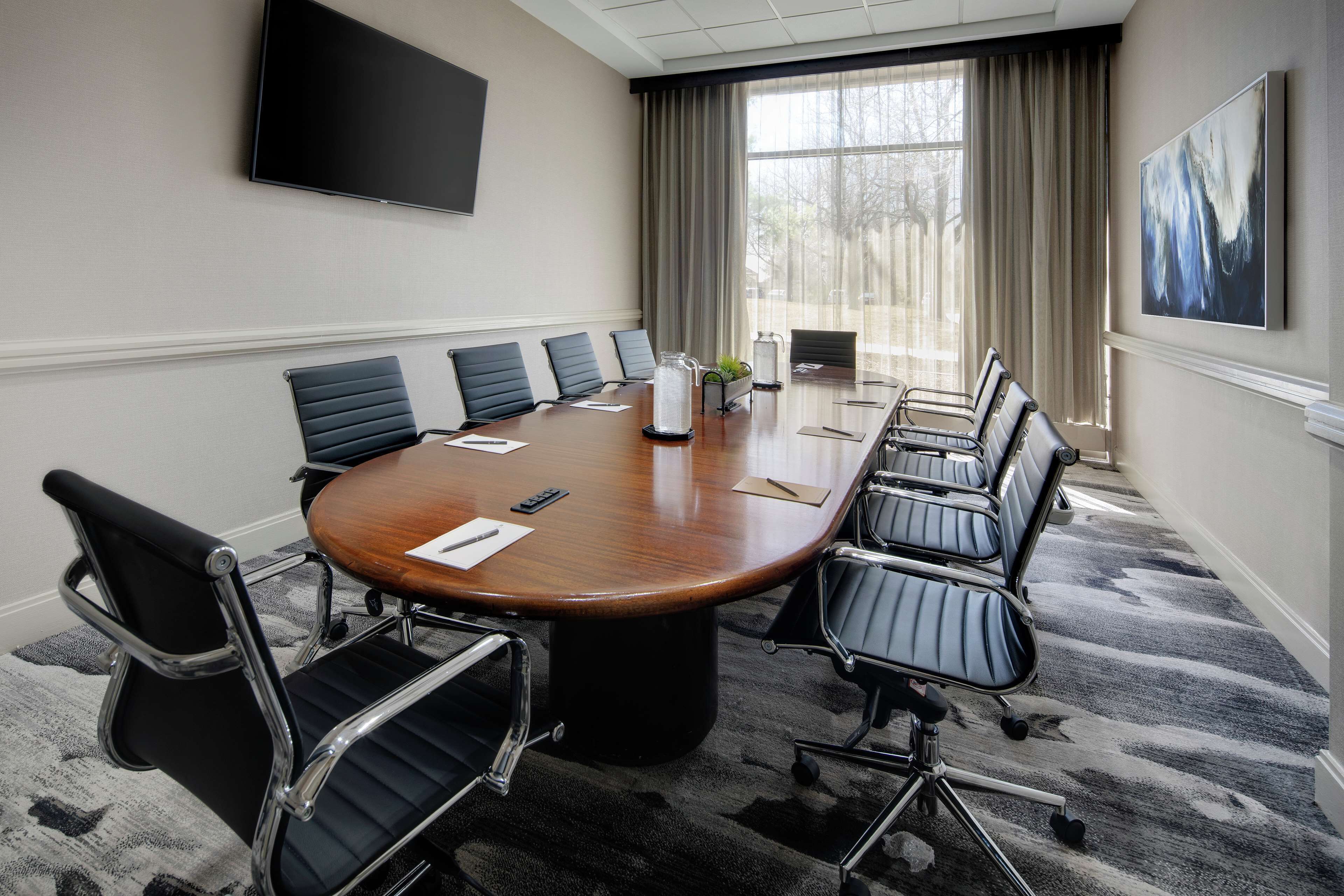 A conference room with a large oval table, black chairs, a flat-screen TV, and a window with curtains.