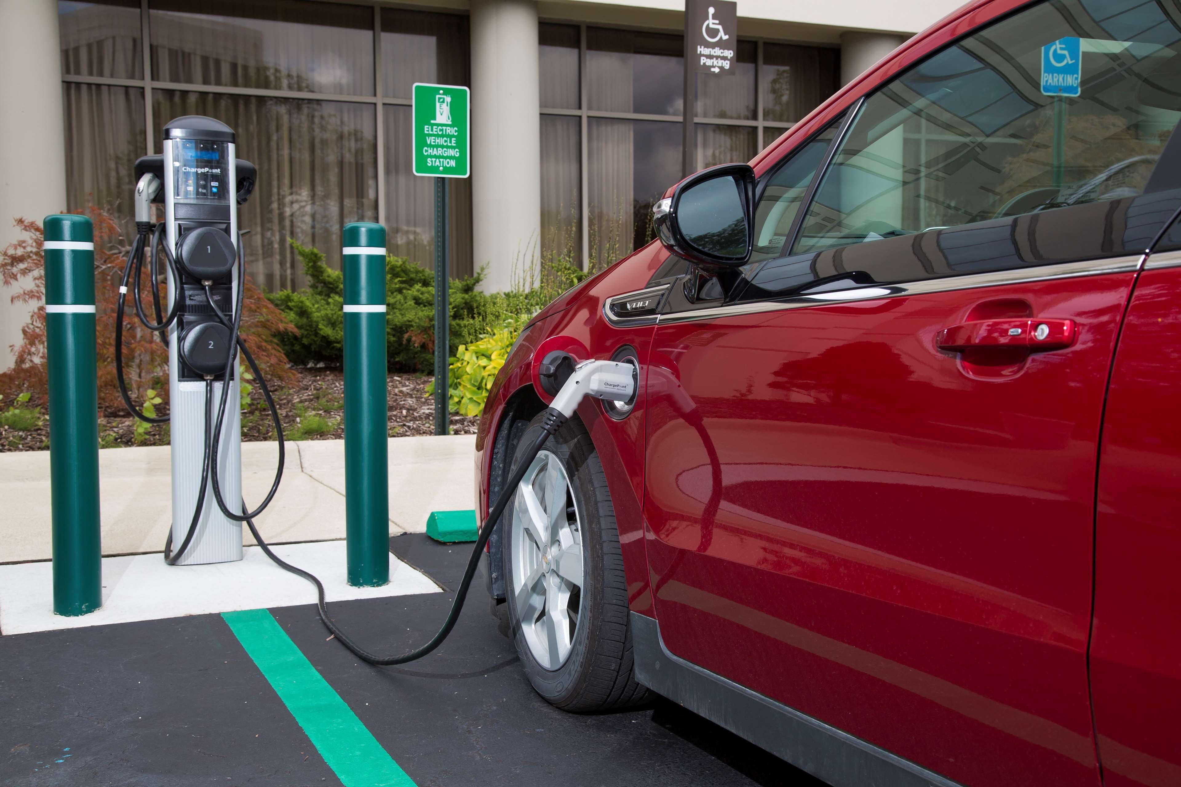 A red car connected to an electric vehicle charging station with a handicap parking sign in the background.
