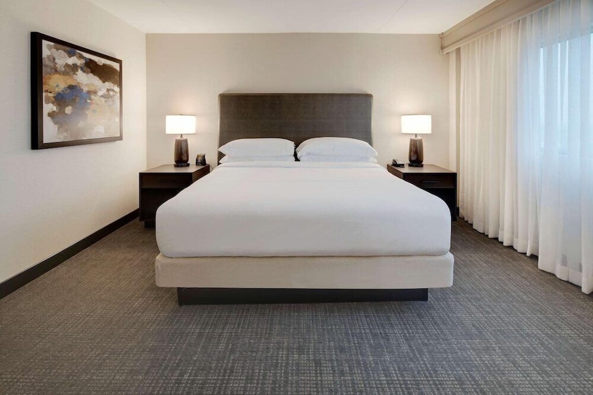 A large bed with a dark headboard, two bedside tables with lamps, a large abstract painting on the wall, and a view of the outside through sheer curtains.