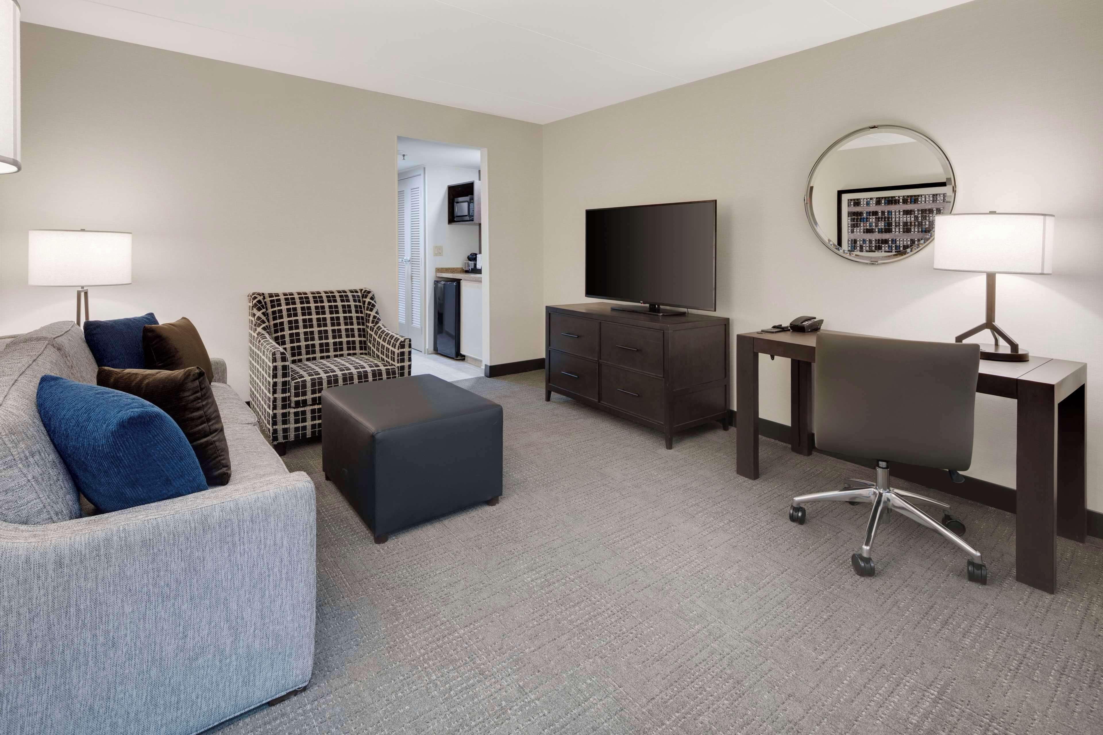 Een hotelkamer met een grijze bank, een gestreept fauteuil, een bureau met een computer, een televisie en een spiegel.