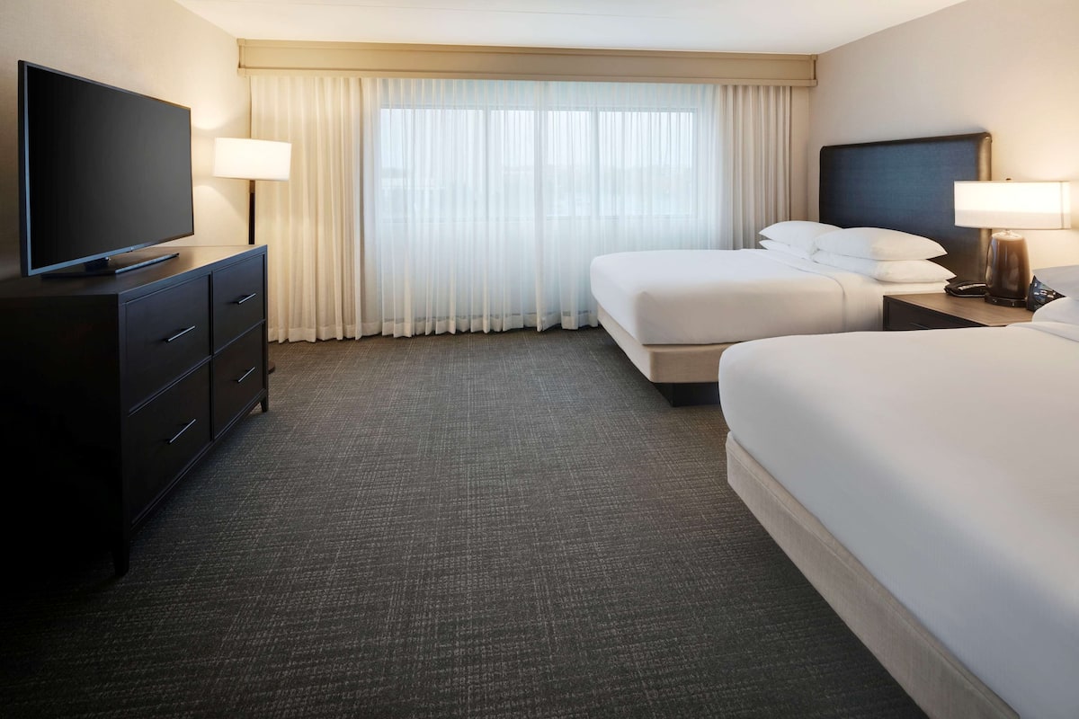 A hotel room with a flat-screen TV on a dresser, two beds, a nightstand with a lamp, and a large window with sheer curtains.