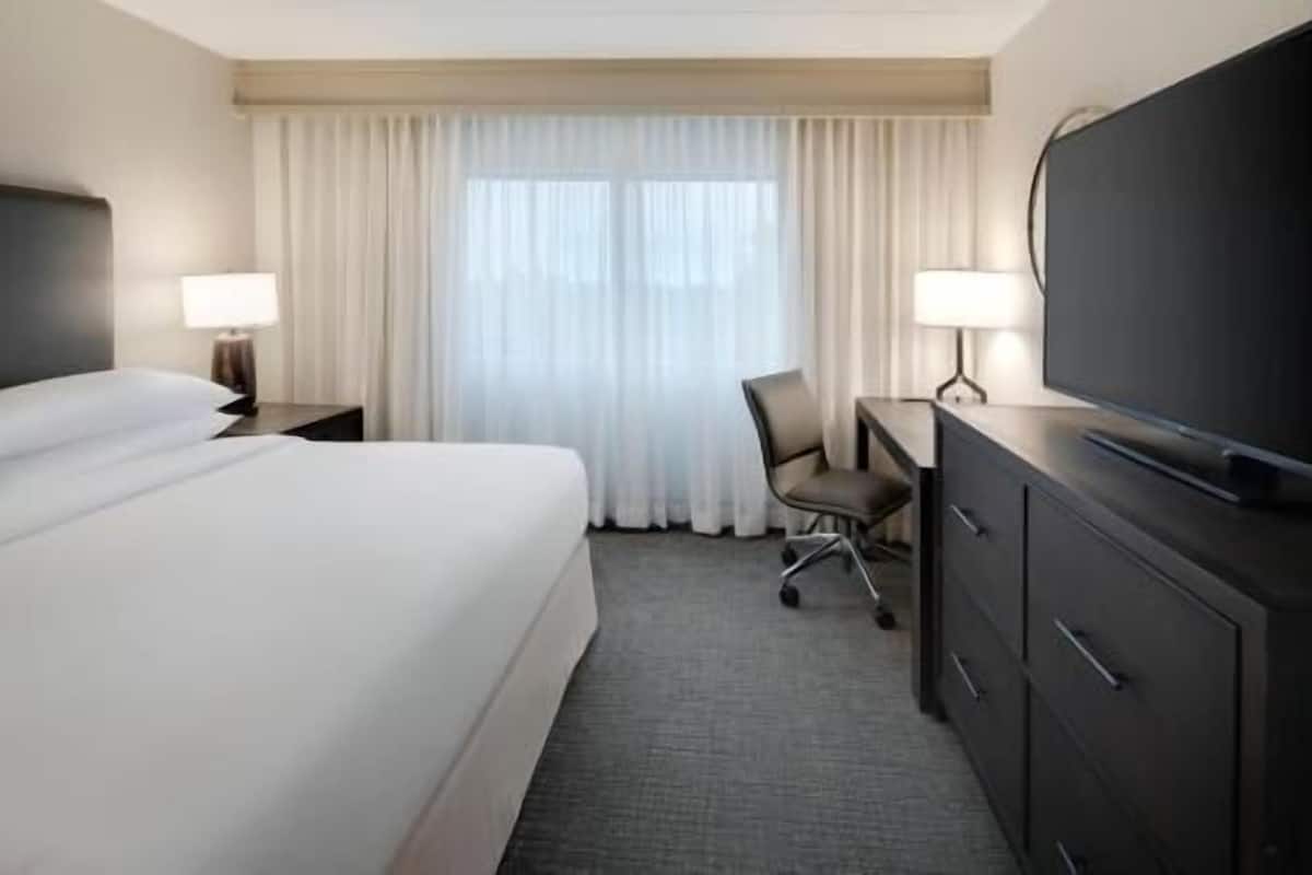 A hotel room with a bed, a desk with a chair, a television, and a window with curtains.