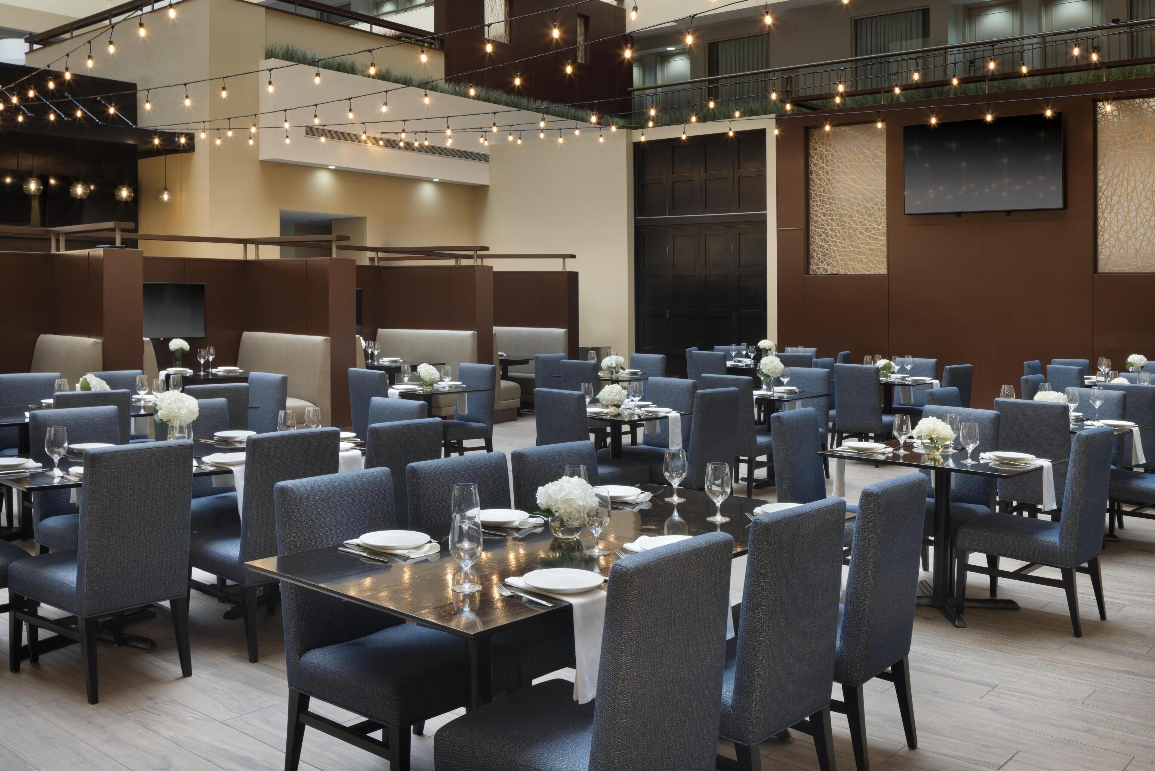 A spacious dining area with multiple tables and chairs, string lights overhead, and a large screen on the wall.