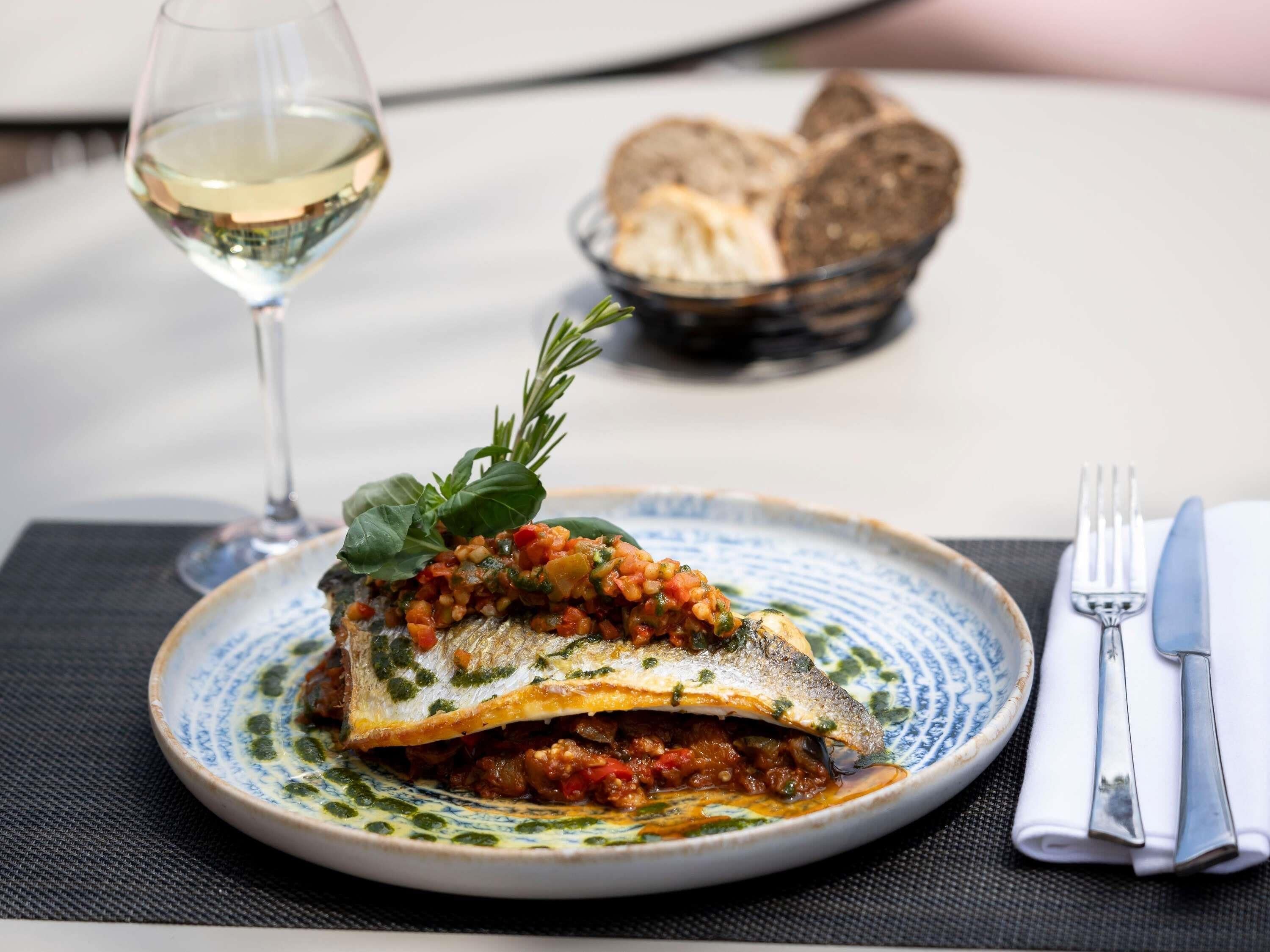 Une assiette de poisson nappée d’une sauce à base de tomates, agrémentée d’herbes, accompagnée de pain et d’un verre de vin blanc, le tout posé sur une table.