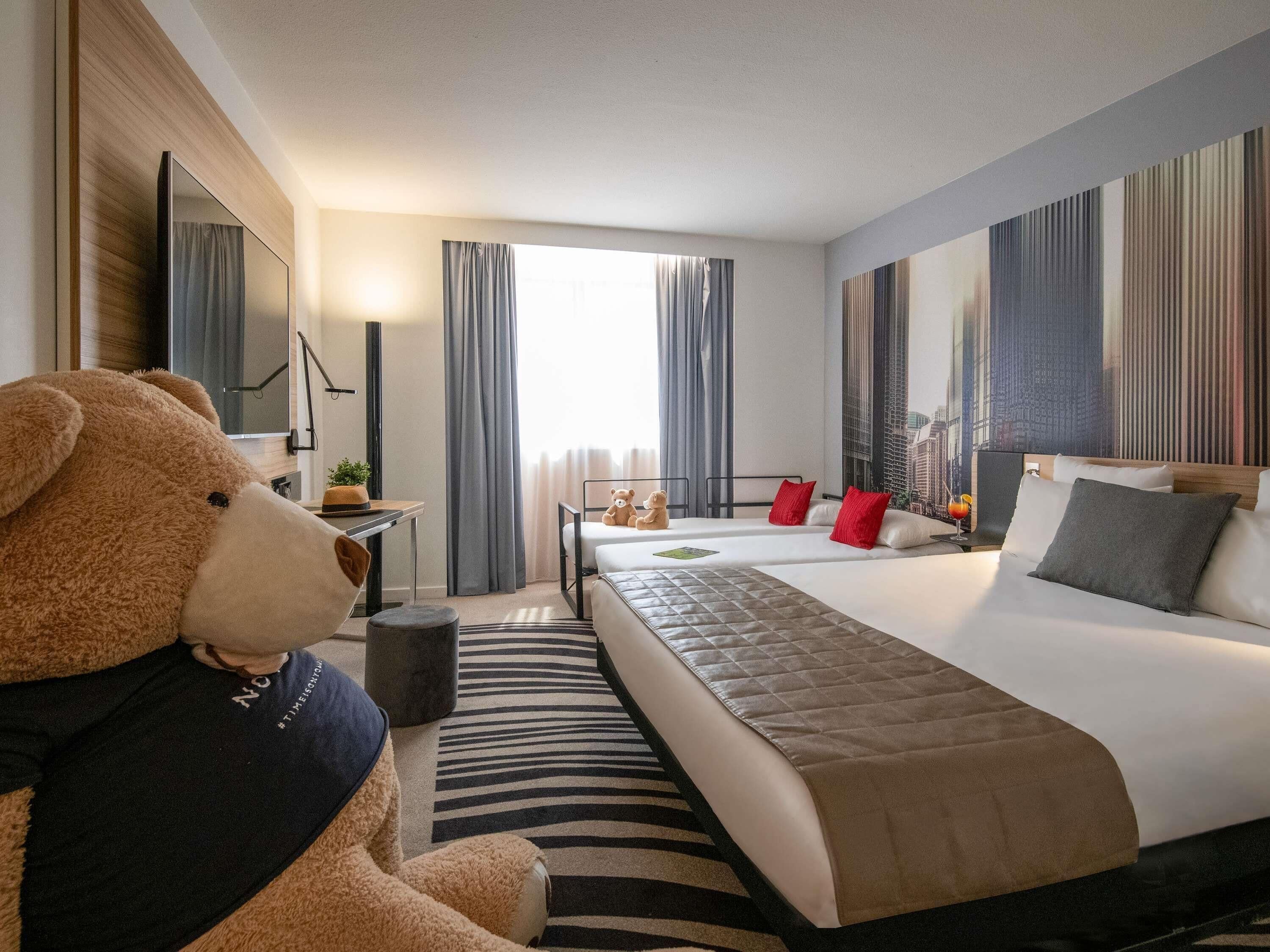 Une chambre d’hôtel avec un grand lit, un bureau, une chaise, un ours en peluche et une grande fenêtre avec des rideaux.