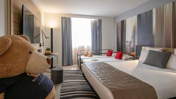 Une chambre d’hôtel avec un grand lit, un bureau, une chaise, un ours en peluche et une grande fenêtre avec des rideaux.