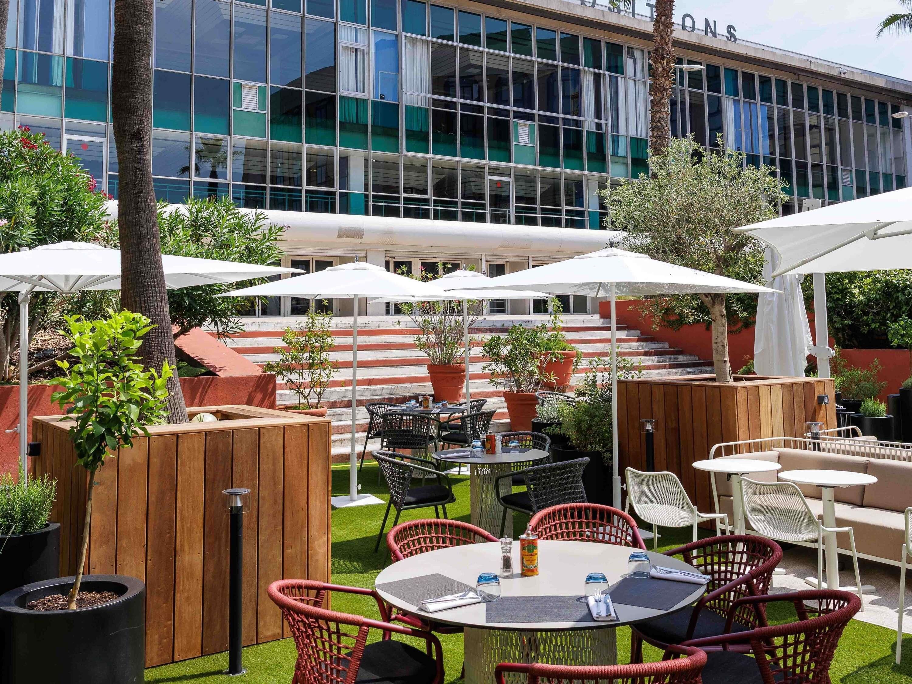 An outdoor seating area with tables and chairs, surrounded by potted plants and umbrellas.