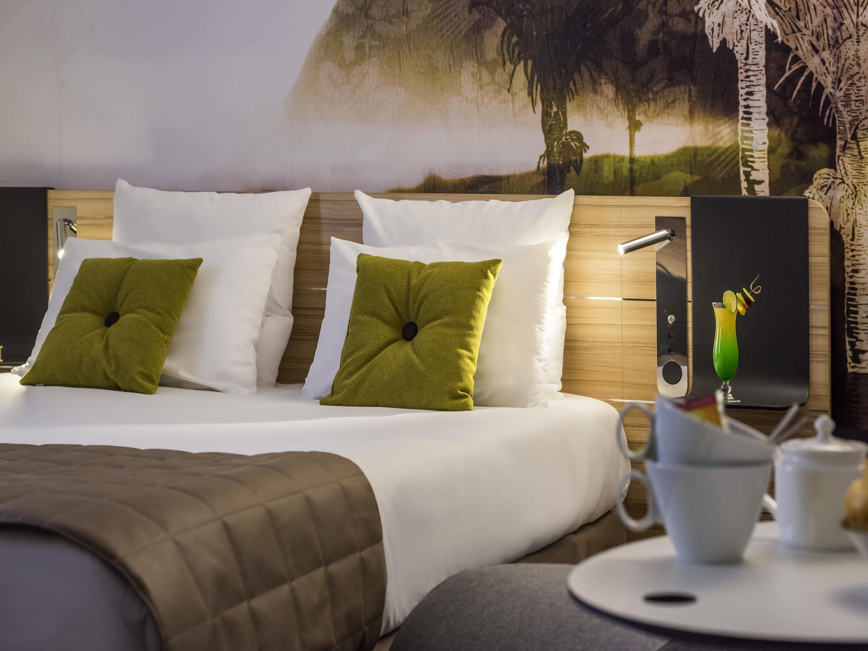 A hotel room with a bed, pillows, a nightstand, a lamp, a painting, and a table with a cup and saucer.