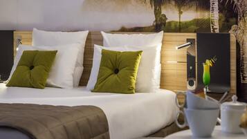 A hotel room with a bed, pillows, a nightstand, a lamp, a painting, and a table with a cup and saucer.