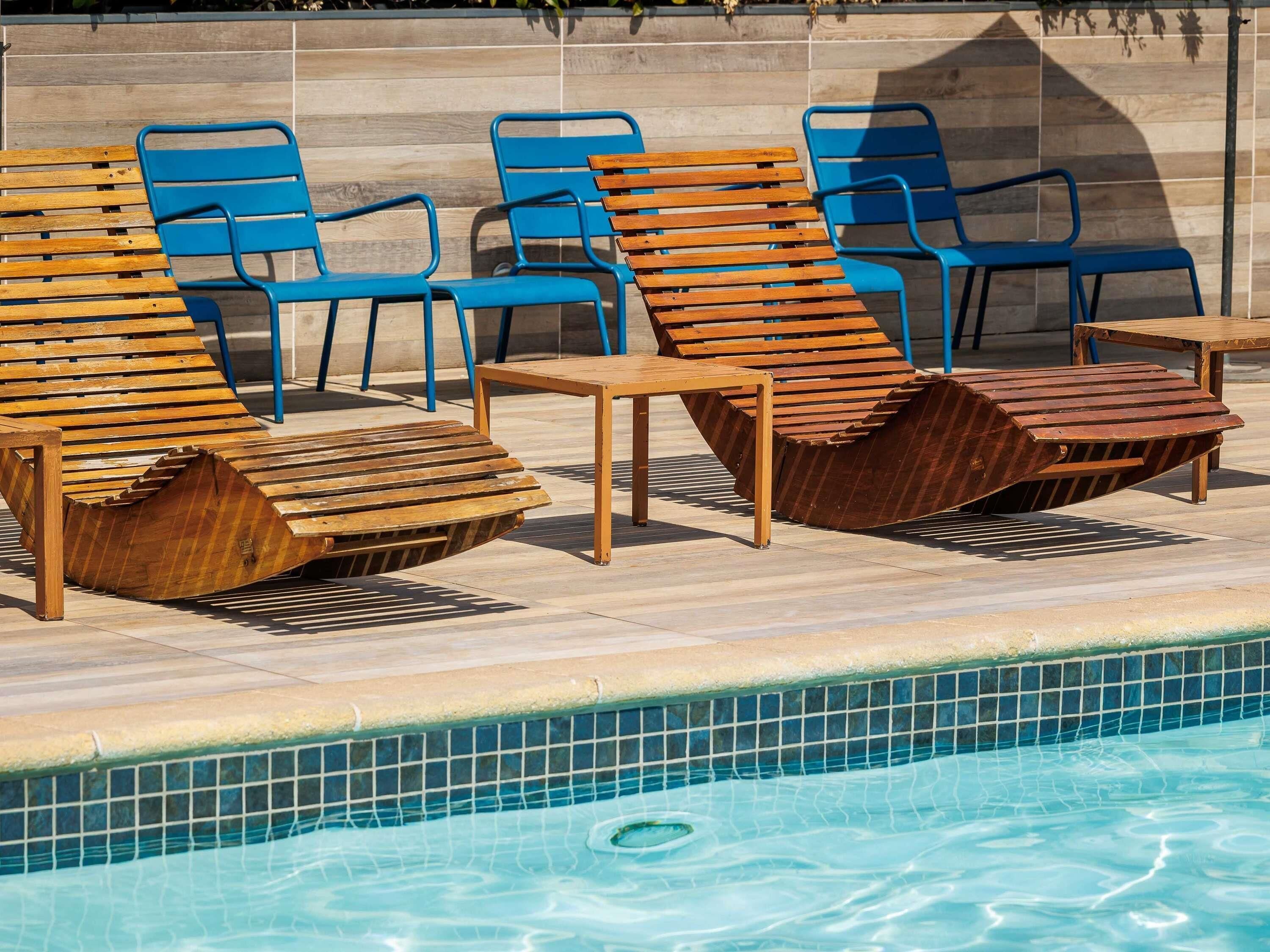 A poolside area with wooden lounge chairs and blue chairs, a small wooden table, and a tiled pool edge.