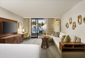 A hotel room with a bed, a TV, a desk, a sofa, a balcony with a view of palm trees and the sea, and decorative wall art.
