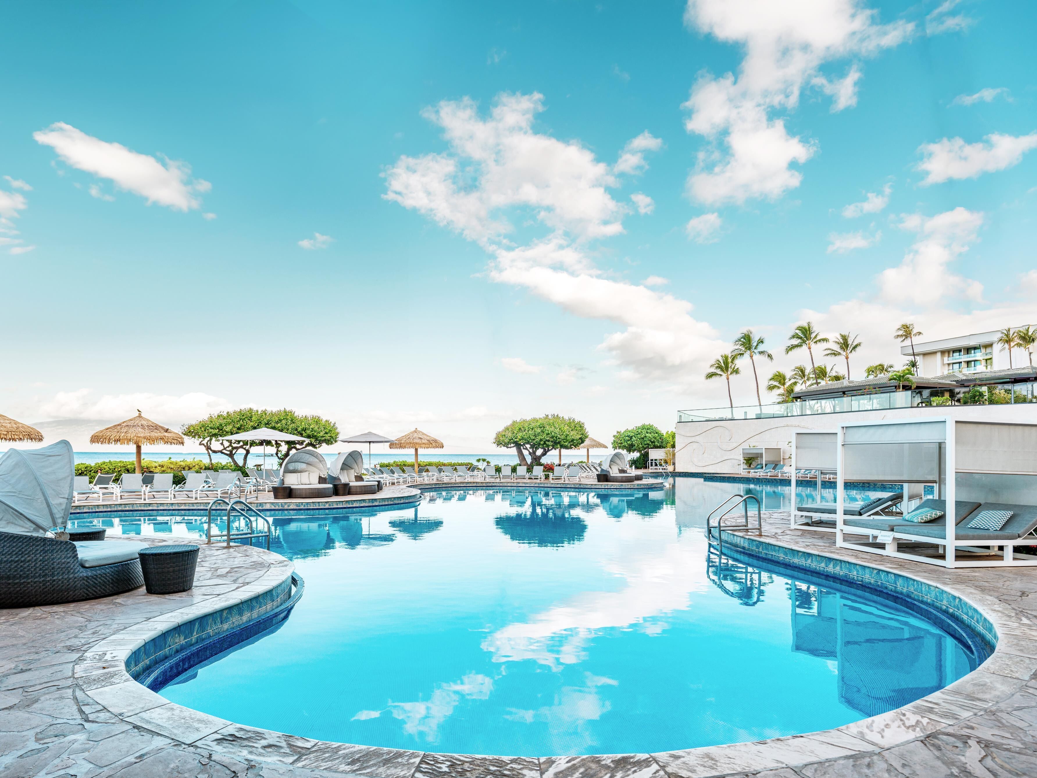 Ein Poolbereich mit Liegestühlen und Sonnenschirmen, umgeben von Palmen und blauem Himmel.