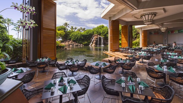 A restaurant with outdoor seating overlooking a pond and waterfall, featuring wicker chairs and tables with green napkins.