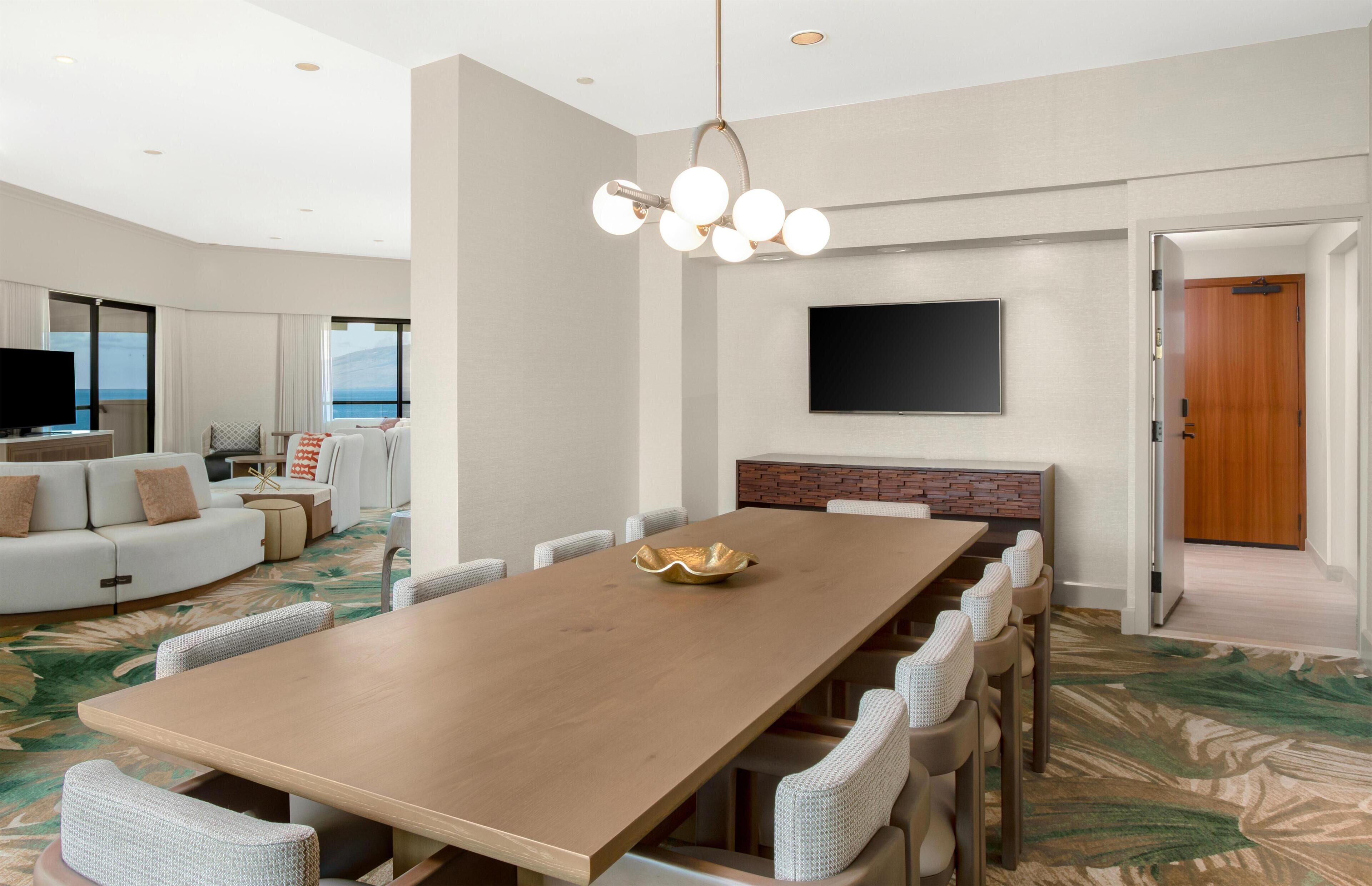 Une chambre d’hôtel moderne dotée d’une grande table à manger, de sièges confortables, d’un téléviseur à écran plat et offrant une vue sur l’océan.