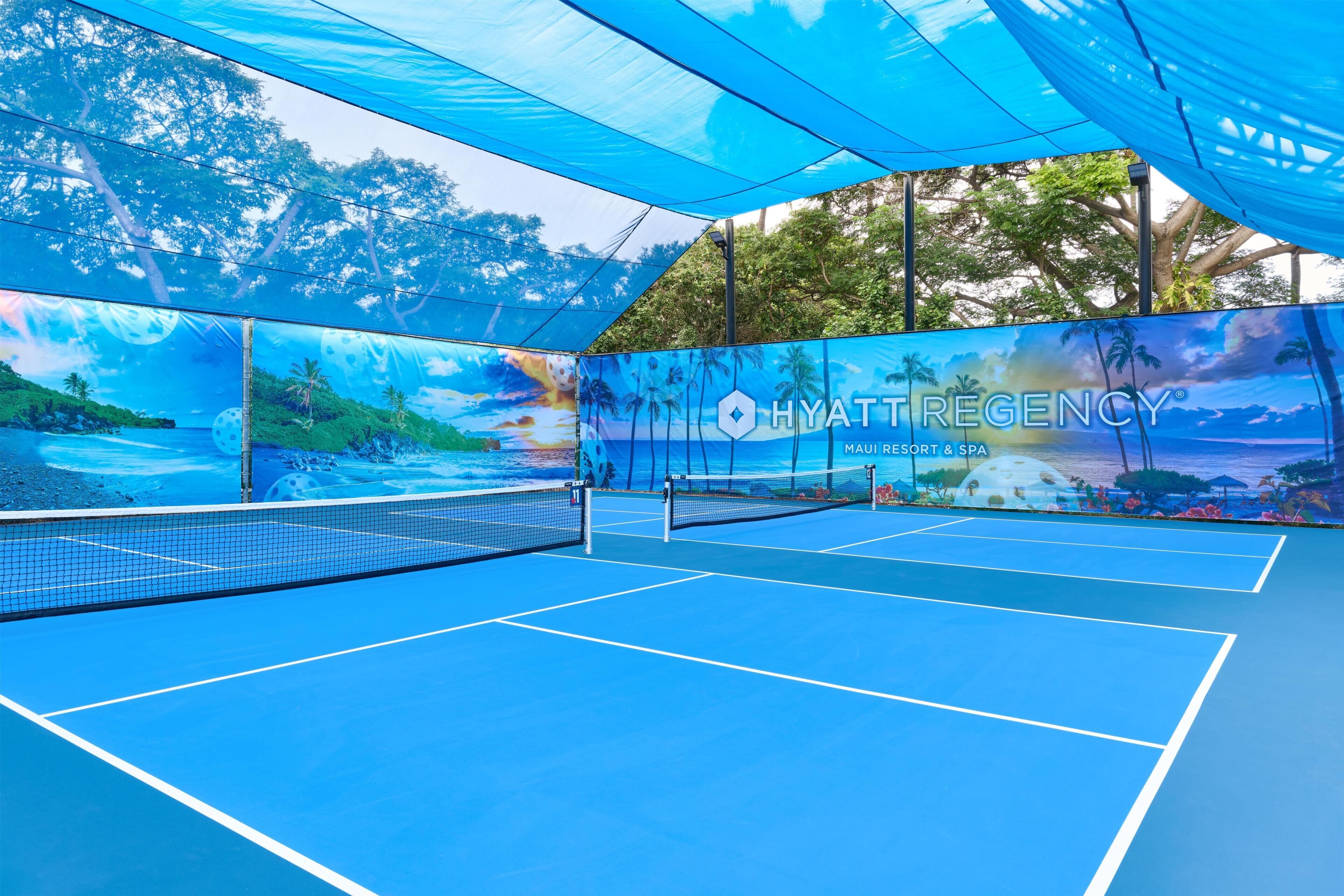 An indoor tennis court with a net, blue flooring, and a large mural depicting a tropical beach scene.