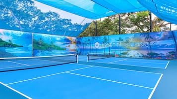 An indoor tennis court with a net, blue flooring, and a large mural depicting a tropical beach scene.