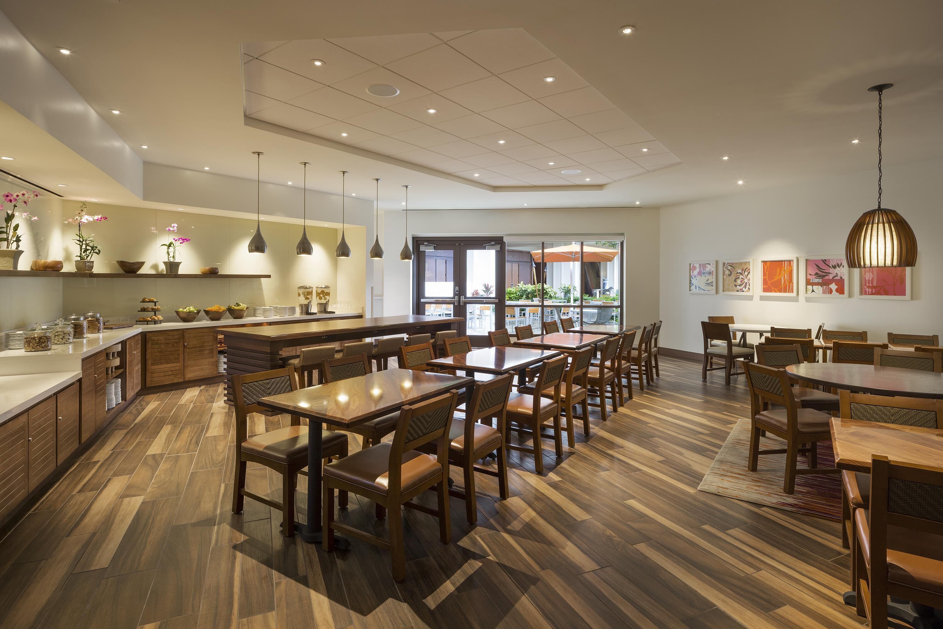 A modern restaurant with wooden flooring, multiple dining tables, and hanging pendant lights.