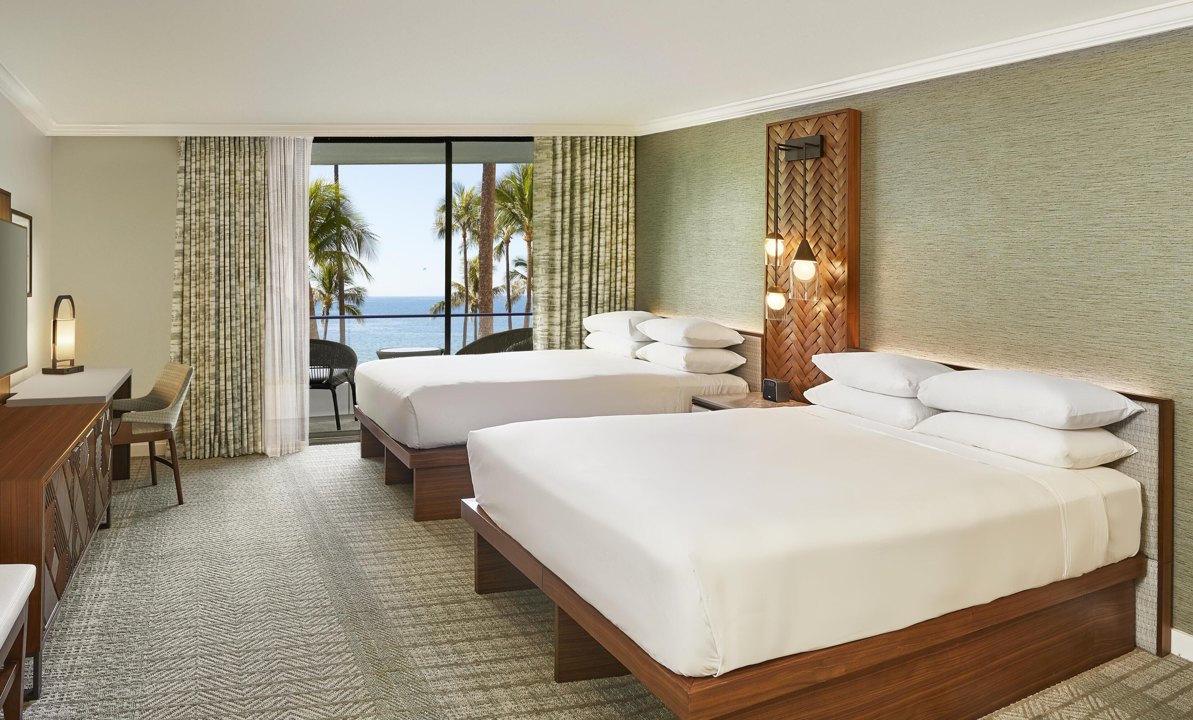 A hotel room with two beds, a desk, a chair, a TV, and a view of the ocean.