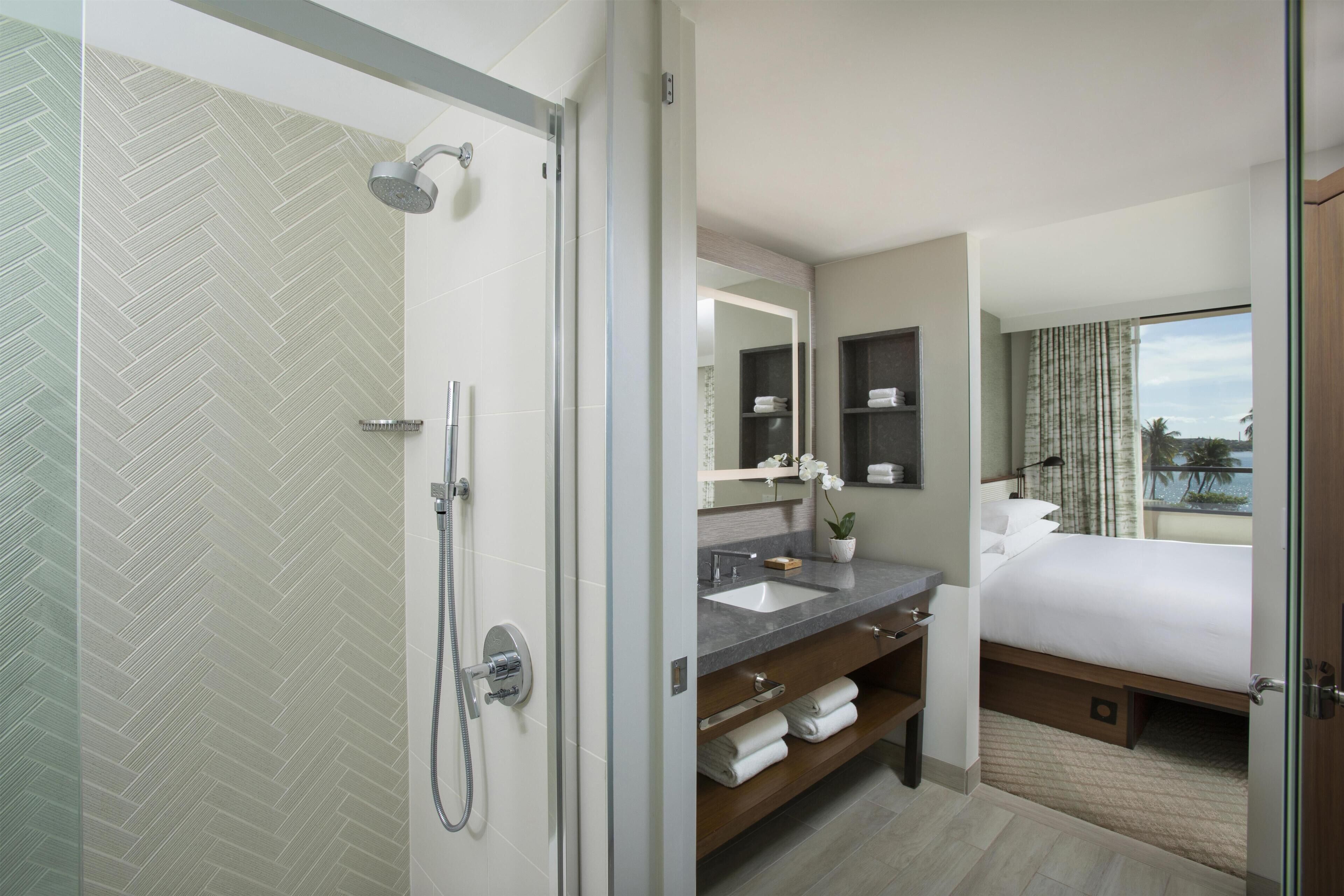 A hotel bathroom with a shower, a sink, a mirror, and a bed with a view.