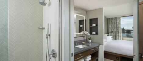 A hotel bathroom with a shower, a sink, a mirror, and a bed with a view.