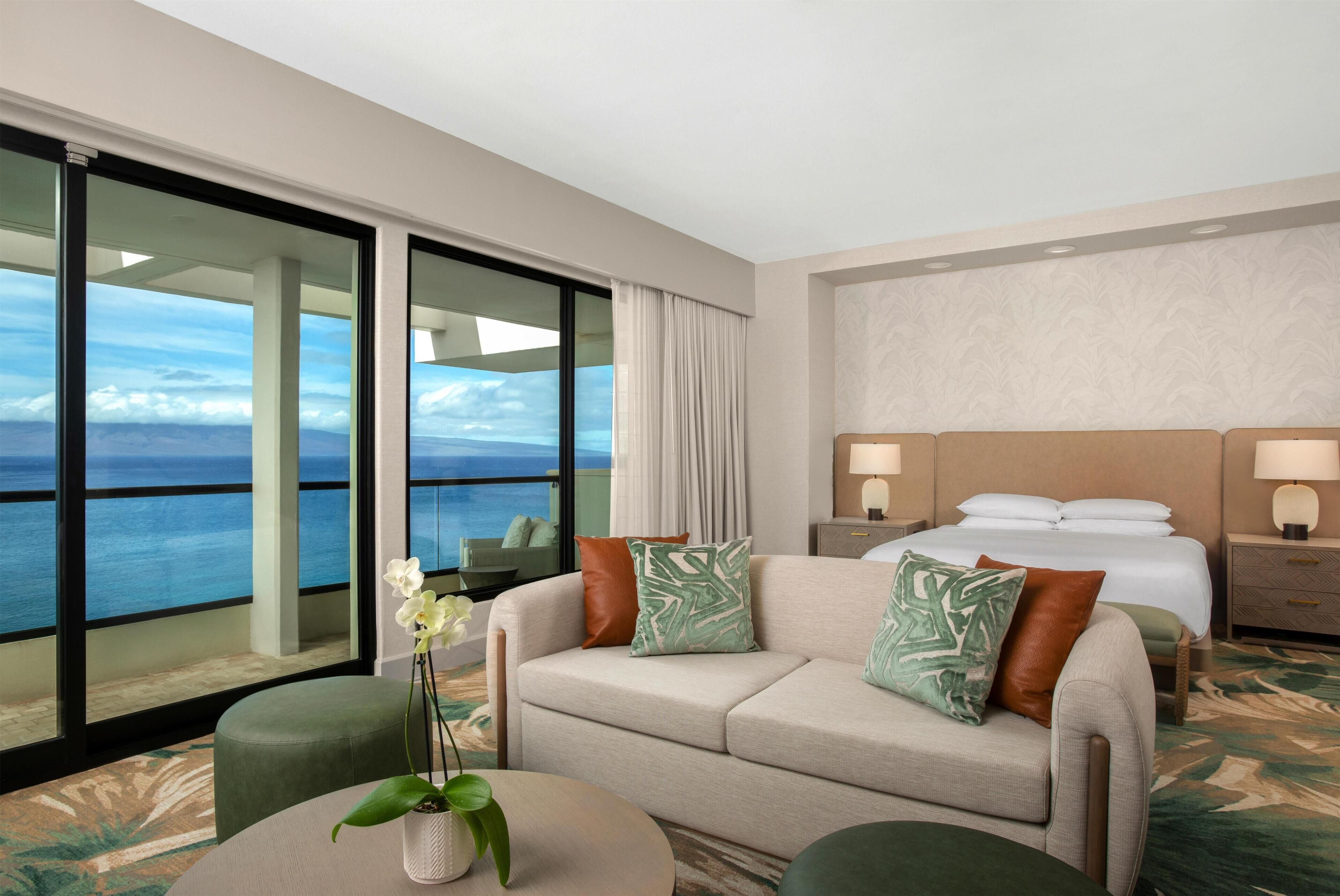 A hotel room with a large sliding glass door leading to a balcony with a view of the sea. There is a bed with white linens, a nightstand with a lamp, and a sofa with decorative pillows.