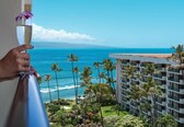 A hand holding a glass with a purple flower, overlooking a coastal view with palm trees and a building.