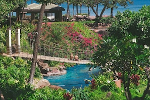 Zona de piscina con un puente, rodeada de exuberante vegetación y vistas al mar.