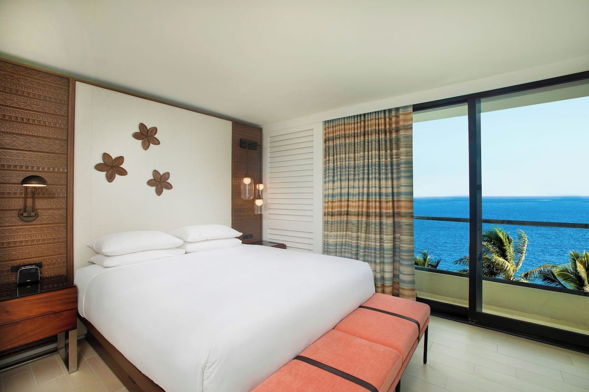 A bedroom with a large bed, a nightstand, a bench, and a view of the ocean.