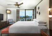 A hotel room with a bed, a TV, a ceiling fan, a balcony with a view of the ocean, and a decorative wall.