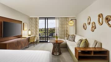 Habitación de hotel con una cama grande, un sofá, un televisor, un escritorio y un balcón con cortinas.