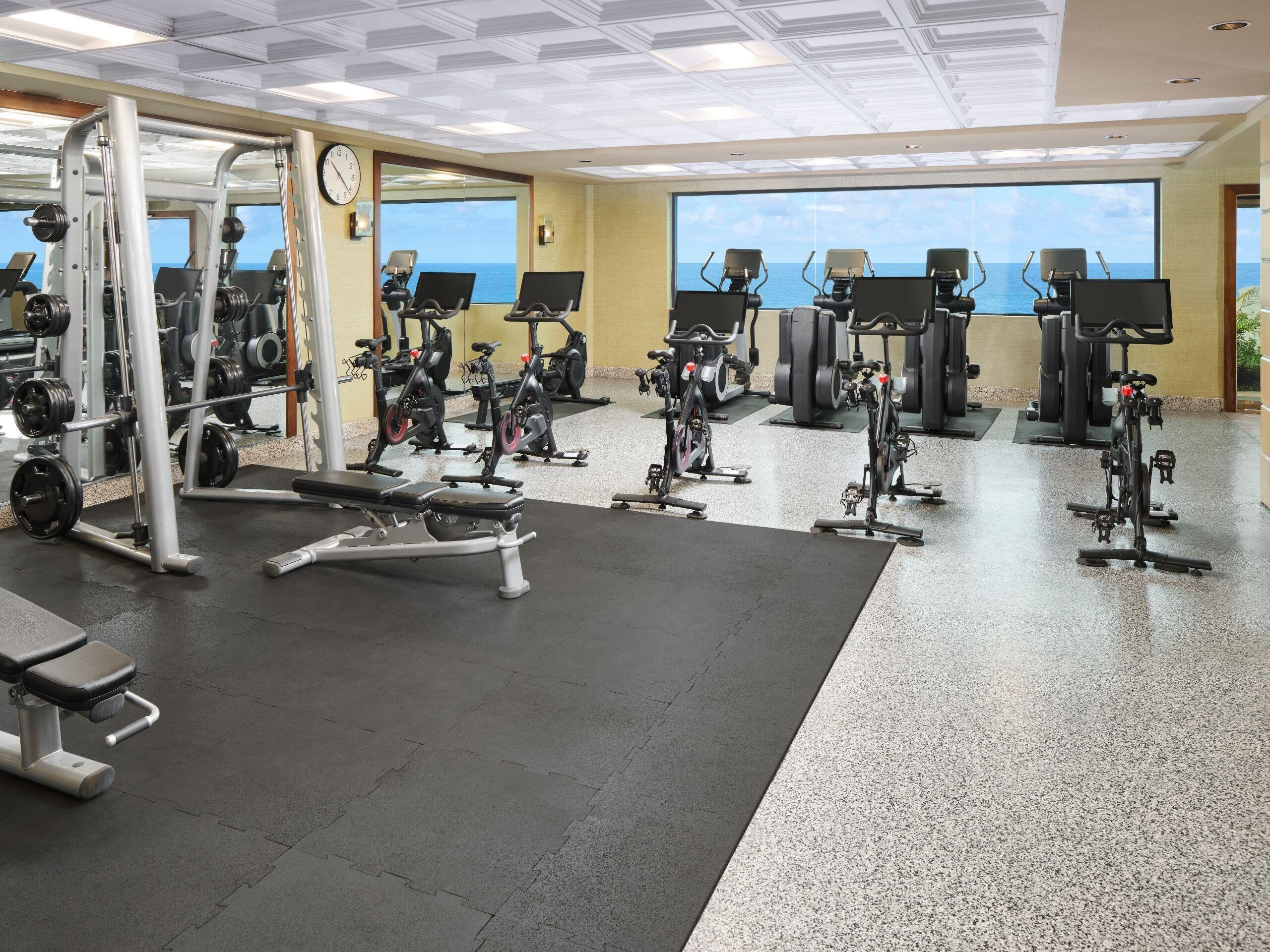 A gym with various exercise equipment, including treadmills and weight machines, and a large window overlooking the sea.