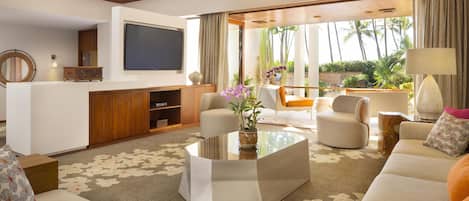 Uma sala de estar moderna com televisão de tela plana, assentos confortáveis, uma mesa de centro e vista para uma área externa tropical.
