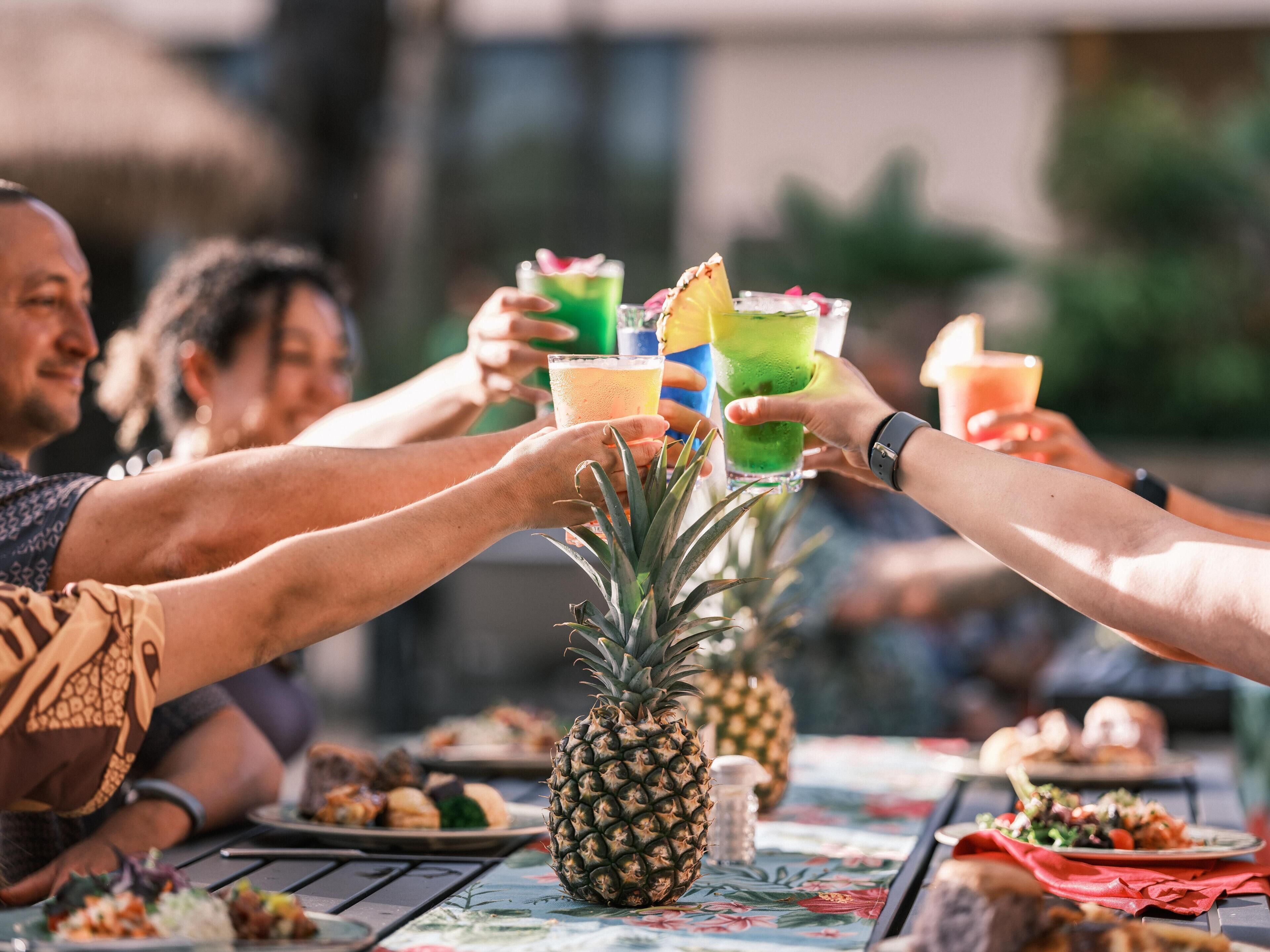 A group of people toasting with colorful drinks at a table with food and a pineapple.