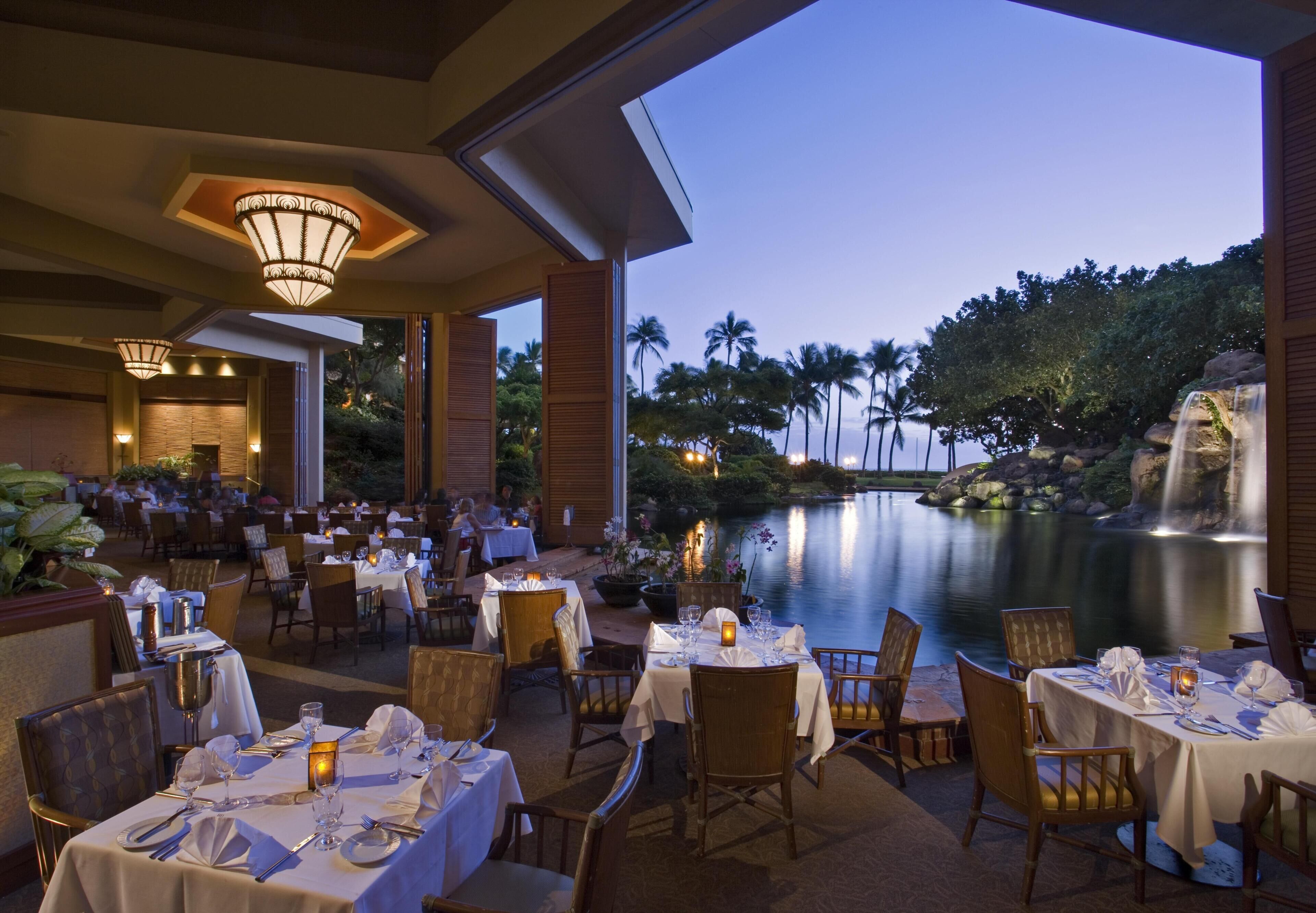 An outdoor dining area with tables set for guests, overlooking a calm body of water and a landscaped garden.