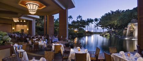 An outdoor dining area with tables set for guests, overlooking a calm body of water and a landscaped garden.