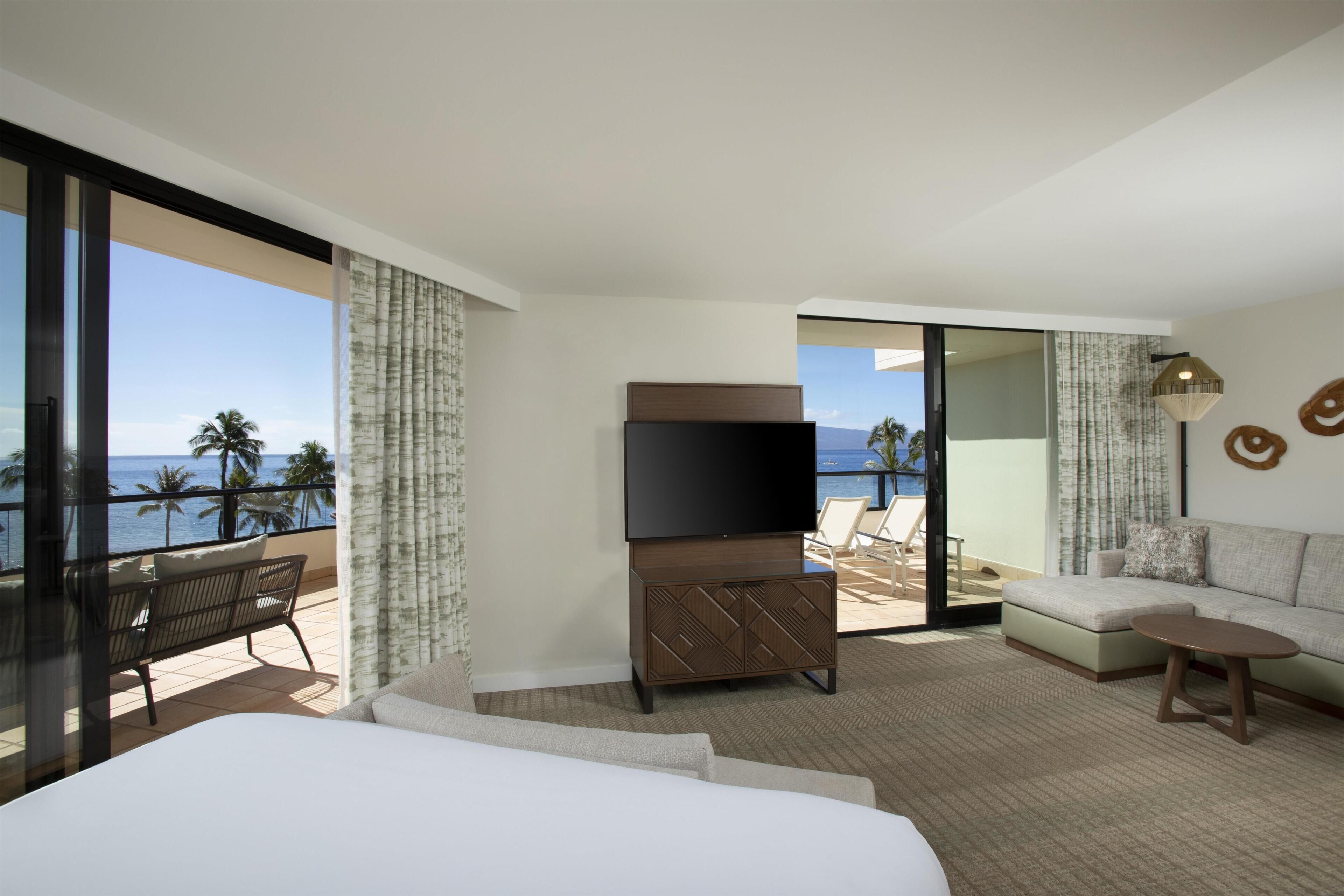 A hotel room with a large bed, a flat-screen TV, a sofa, and a balcony with a view of the ocean.