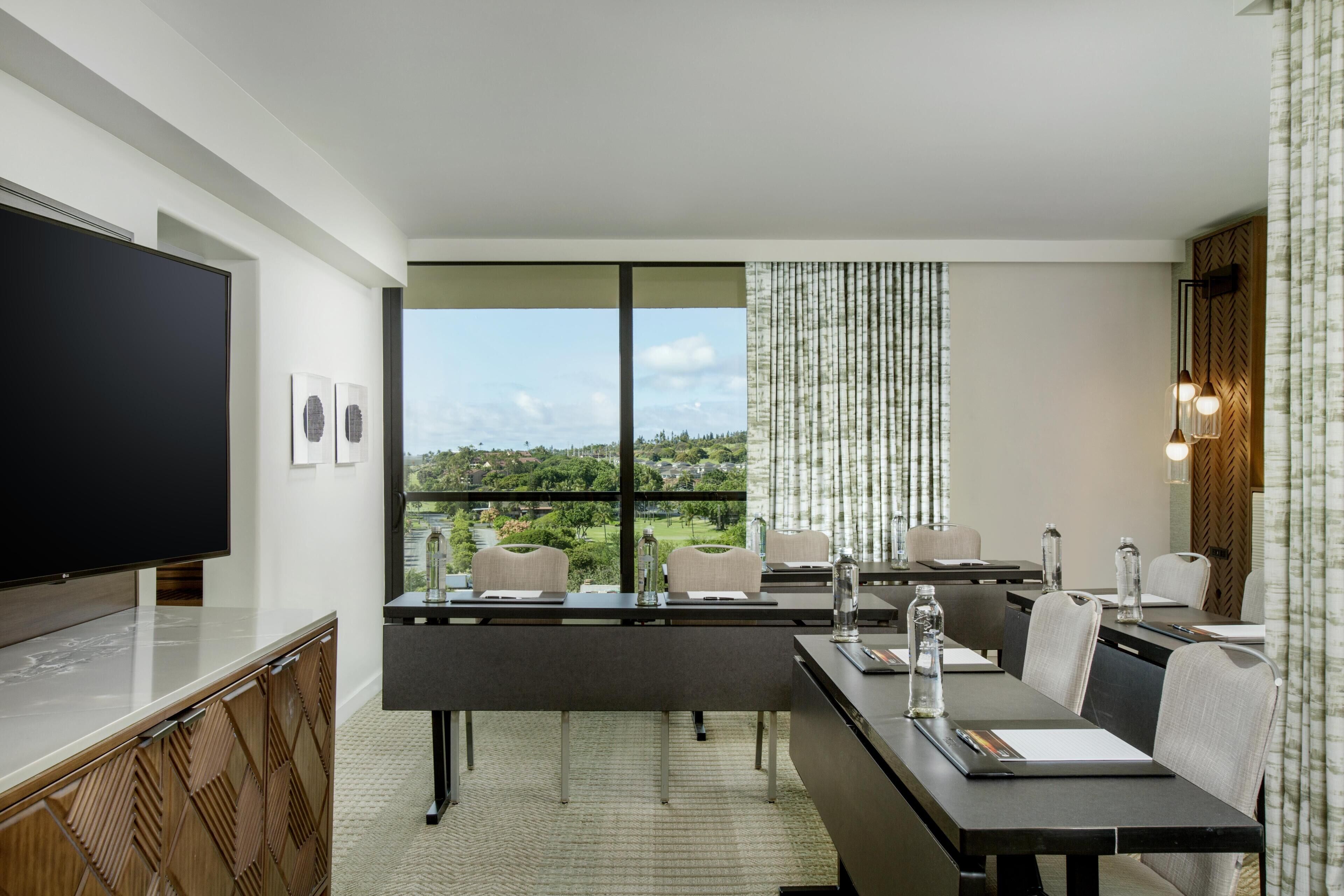 Ein modernes Hotelzimmer mit einem großen Flachbildfernseher, einem Essbereich mit Tisch und Stühlen sowie Blick auf eine grüne Landschaft durch die Fenster.
