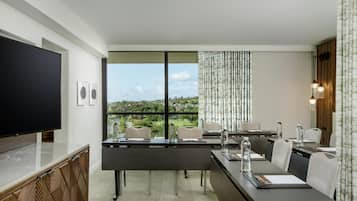 Une chambre d’hôtel moderne dotée d’une grande télévision à écran plat, d’un coin repas avec une table et des chaises, et offrant une vue sur un paysage verdoyant par les fenêtres.