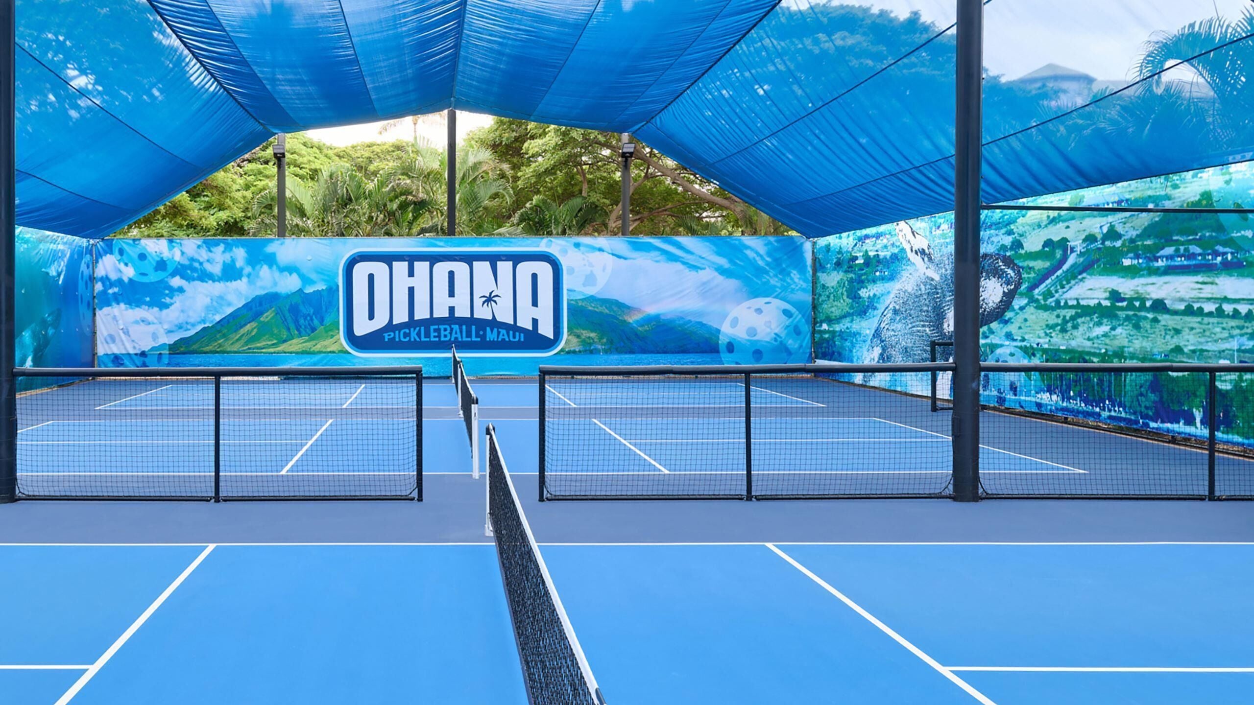 An indoor tennis court with a net, blue flooring, and a large banner with 'OHANA PICKLEBALL MUI' text.