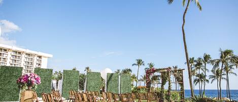 An outdoor wedding setup with chairs arranged on grass, a backdrop of greenery, and a view of the ocean.